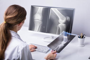 In a hospital setting, a radiographer prepares a patient for an X-ray while a radiologist reviews CT scans on a monitor.